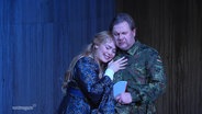 Auf einer Theaterbühne legt eine Schauspielerin ihren Kopf auf die Brust eines Mannes, der eine Bundeswehr-Uniform trägt. © Screenshot 