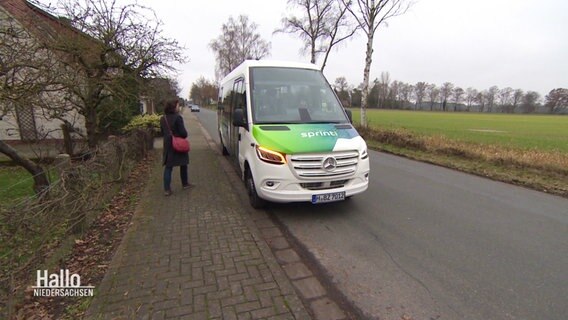 Eine Frau wartet am Straßenrand auf den gerade ankommenden Rufbus. © Screenshot 