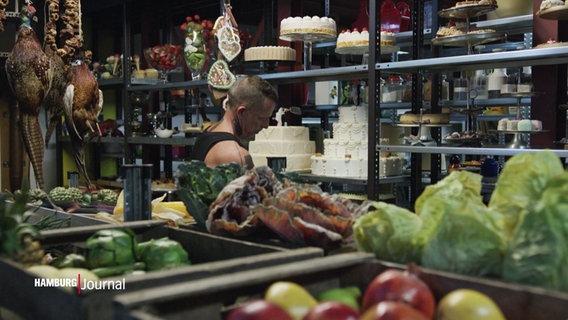 Eine Person steht zwischen Regalen voll künstlich nachgebildeter Lebensmittel wie Torten, Gemüse, Salat oder Kuchen. © Screenshot 