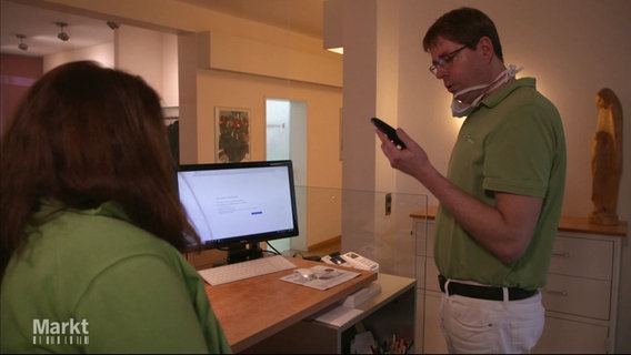 Im Empfangsbereich einer Zahnarztpraxis: Ein Mann schaut auf ein Telefon in seiner Hand, der Bildschirm eines Computers zeigt an, dass keine Seite geladen werden kann. © Screenshot 