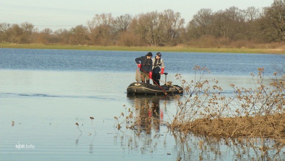 Zwei Personen mit Keschern in der Hand stehen in einem Schlauchboot, das auf einem überfluteten Gebiet treibt. © Screenshot 