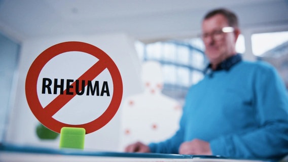 Im Vordergrund ein Schild mit der rot durchgestrichenen Aufschrift "Rheuma", im Hintergrund unscharf zu sehen eine Person. © Screenshot 