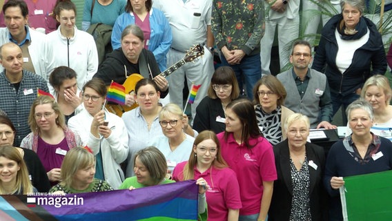 Eine Gruppe von Menschen steht beisammen mit Regenbogenflaggen und Schildern gegen rechts. © Screenshot 