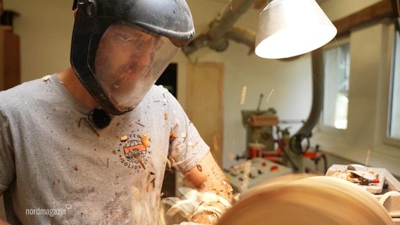 Ein Mann verarbeitet Holz und trägt dabei einen Gesichtsschutz. © Screenshot 