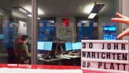 Im Vordergrund rechts unten eine Texttafel mit der Aufschrift: "30 Johr Nachrichten op platt". Im Hintergrund ein Büro mit mehreren Bildschirmen und Schreibtischen. © Screenshot 