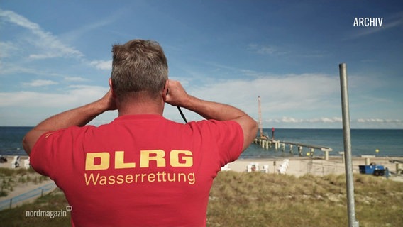 Rückenansicht eines Mannes, der ein rotes DLRG T-Shirt trägt und durch ein Fernglas auf das Meer schaut. © Screenshot 