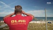 Rückenansicht eines Mannes, der ein rotes DLRG T-Shirt trägt und durch ein Fernglas auf das Meer schaut. © Screenshot 