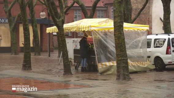 Eine Person steht an einem Marktstand unter einer Plane, geschützt vom Regen. © Screenshot 