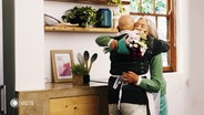 Eine ältere Dame und ein älterer Herr umarmen sich. Sie hält dabei einen Blumenstrauß in der Hand. © Screenshot 
