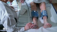 Ein älterer Mann liegt auf einer Liege und wird von einer Ärztin an den Beinen untersucht. © Screenshot 