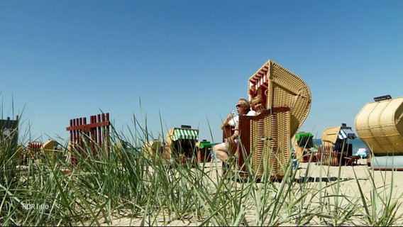 Menschen sitzen in Strandkörben am Meer und genießen die Sonne. © Screenshot 