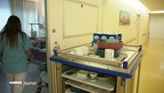 Eine Pflegekraft betritt ein Zimmer in einem Krankenhaus, von hinten zu sehen. Neben der Tür steht ein Rollwagen mit Bechern und Schüsseln. © Screenshot 
