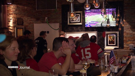 In einer Kneipe in Hamburg während des Super Bowl vom American Football.: Fans in roten Trikots schauen auf einen Fernseher, am Tresen sieht man eine kleine USA-Flagge. © Screenshot 
