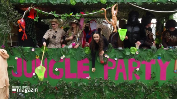 Ein grün geschmückter Karnevalswagen, aufdem "Jungle Party" steht und von dem kostümierte Karnevalisten Süßigkeiten oder ähnliches werfen. © Screenshot 