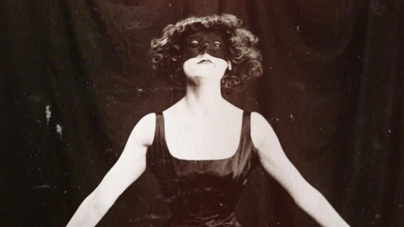 Asta Nielsen steht in einem dunklen Trägeroberteil und einer dunklen Maske über den Augen vor einem Vorhang. © Screenshot 