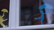 Eine Fensterscheibe in der Fensterbilder von Kindern mit Regenschrimen zu sehen sind. © Screenshot 