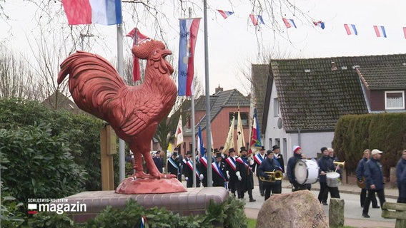 Ein Spielmannszug des Heimatvereins Südereggen zieht über eine Straße. Im Vordergrund ist eine rote Statue von einem Hahn. © Screenshot 