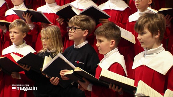 Ein Junge in schwarzer Kleidung, steht in der Mitte von weiteren Jungen, die bereits die roten Roben des Kieler Knabenchors tragen. © Screenshot 