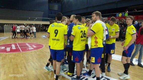 HC Empor Rostock steht nach einem Spiel zusammen im Kreis in der Handballhalle. Im Hintergrund jubelt MTV Braunschweig. © Screenshot 