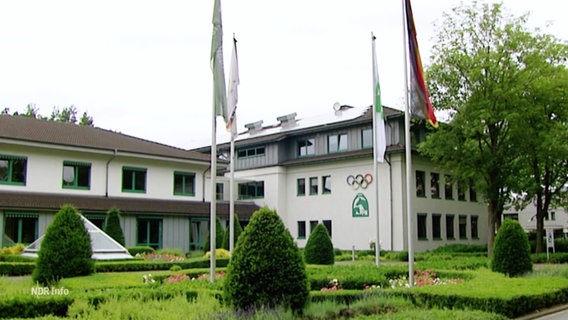 Auf einem Gebäude sind ein Logo mit zwei Pferdeköpfen sowie die olympischen Ringe zu sehen. © Screenshot 