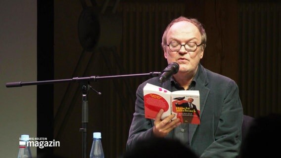 Jens Wawrczeck sitzt auf einer Bühne und liest aus seinem Buch vor. © Screenshot 