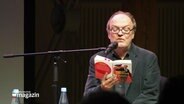 Jens Wawrczeck sitzt auf einer Bühne und liest aus seinem Buch vor. © Screenshot 