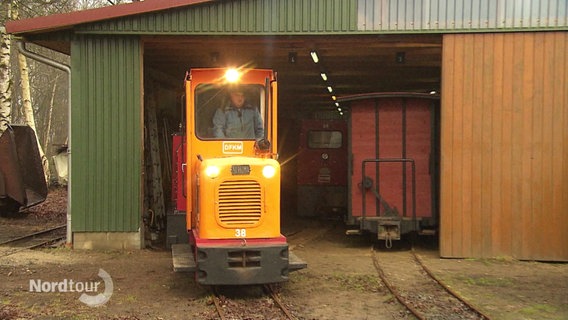 Die orangefarbene Lok einer Kleinbahn fährt aus einem Schuppen. © Screenshot 