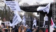 Die Hamburger Band Deichkind spielt auf einer Demo gegen Rechtsextremismus am 25.02.2024. Sie tragen weiße Anzüge und schwenken weiße Fahnen, auf beidem steht etwas geschrieben. © Screenshot 