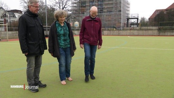 Drei Menschen stehen auf einem Sportfeld. Die Frau in der Mitte trägt keine Schuhe. © Screenshot 