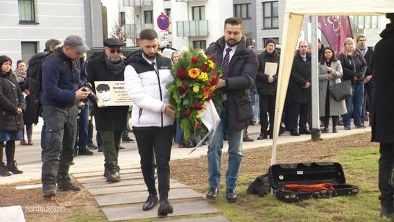 Menschen bei einer Gedenkveranstaltung für Mehmet Turgut am 25.02.2024. Im Vordergrund tragen zwei Männer einen Trauerstrauß. © Screenshot 