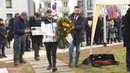 Menschen bei einer Gedenkveranstaltung für Mehmet Turgut am 25.02.2024. Im Vordergrund tragen zwei Männer einen Trauerstrauß. © Screenshot 