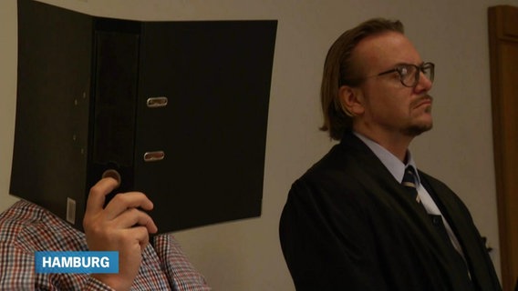 Ein Mann hält einen Aktenordner, um sein Gesicht zu verbergen, neben ihm sein Anwalt. © Screenshot 