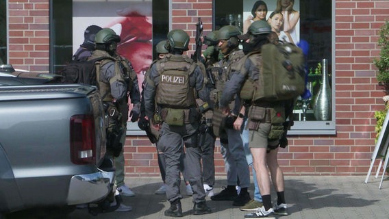 Maskierte und durch ihre Ausrüstung gut geschützte Spezialeinsatzkräfte der Polizei stehen in einer kleiner Gruppen auf dem Bürgersteig. © Screenshot 