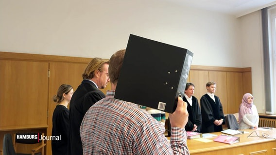 Vor Gericht hält sich ein Mann einen Aktenordner vors Gesicht. © Screenshot 