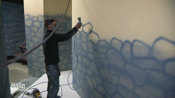 Ein Mann malt mittels Airbrush eine graue Steinmauer an eine Innenwand. © Screenshot 