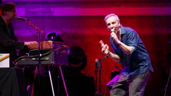 Olli Schulz singt mit Mikrofon in der Hand auf einer Bühne, im Hintergrund spielt ein Keyboarder. © Screenshot 