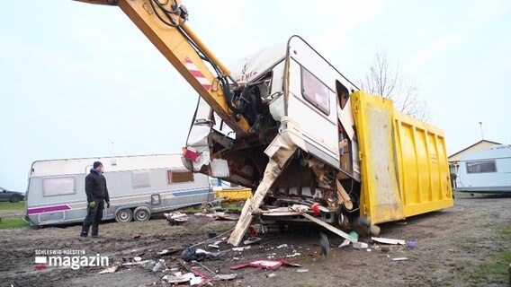 Ein Wohnwagen wird von einem Bagger in einen Container geschoben. © Screenshot 