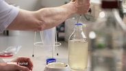 Szene in einem Labor: Eine person entnimmt mit einer Pipette eine klare Flüssigkeit aus einer Flasche. Neben der Flasche liegt eine Petrischale. © Screenshot 