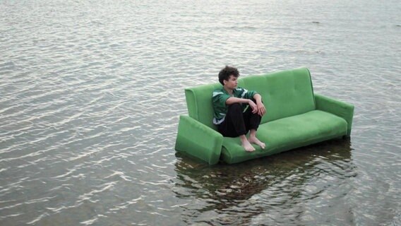 Ein Junge sitzt mit angewinkelten Beinen auf einer grünen Couch, die in einem Gewässer steht. © Screenshot 