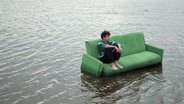 Ein Junge sitzt mit angewinkelten Beinen auf einer grünen Couch, die in einem Gewässer steht. © Screenshot 