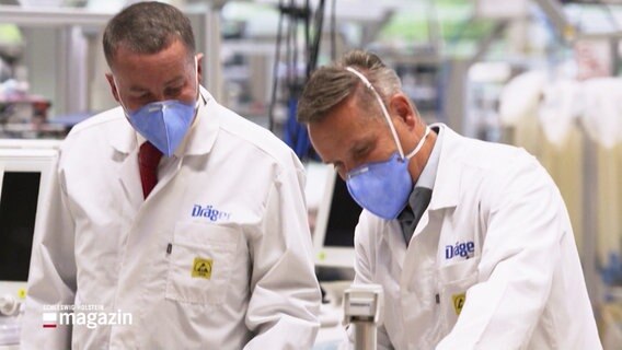Zwei Mitarbeiter in weißen Kitteln und mit Atemschutzmasken im Drägerwerk © Screenshot 