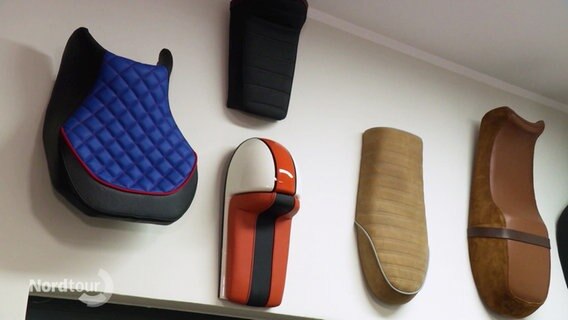 An einer Wand hängen Arbeiten eines Sattlers: Lederbezogene Sitze für Roller, Vespas oder Ähnliches. © Screenshot 