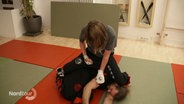 Eine Szene bei der chinesischen Kampfsportart Wing Tsun: In einem Trainingsraum mit Matten auf dem Boden sitzt eine weibliche auf einer männlichen Person und fixiert ihn am Boden. © Screenshot 