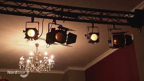 Vier Scheinwerfer hängen an einer schwarzen Traverse, im Hintergrund hängt ein Kronleuchter von der Decke. © Screenshot 