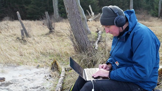 Christian Löffler sitzt auf einem Baumstamm, er trägt Kopfhörer und blickt auf einen Laptop. © Screenshot 