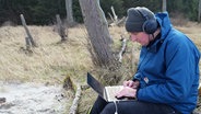 Christian Löffler sitzt auf einem Baumstamm, er trägt Kopfhörer und blickt auf einen Laptop. © Screenshot 