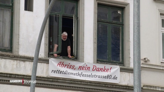 Ein Mann schaut aus dem Fenster eines Hauses, unter dem Fenster hängt ein Banner mit der Aufschrift: "Abriss, nein danke! rettetdiemethfesselstrasse80.de" © Screenshot 