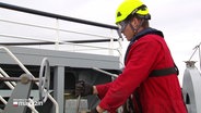 Ein junger Mann in rotem Arbeitsoverall, Sicherheitsbrille, neongelbem Helm und Arbeitshandschuhen bei der Arbeit im Freien auf einem Schiff. © Screenshot 