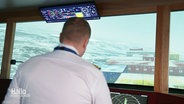 Ein Kapitän steht vor einem großen Screen, der eine Schifffahrt simuliert. © Screenshot 