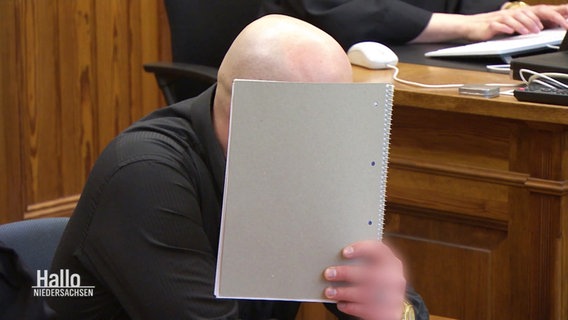 Ein Angeklagter hält sich vor Gericht eine Kladde vor das Gesicht. © Screenshot 